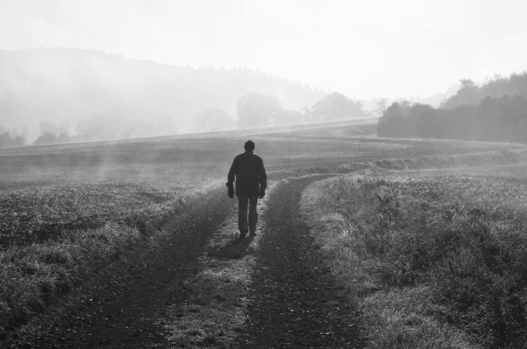 A lone person walks down a foggy rural path, captured in a dramatic black and white scene.
