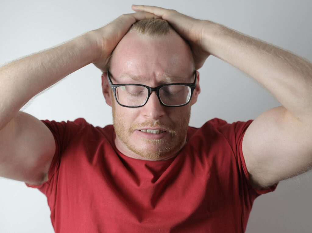 Irritated bearded man in red tee shirt and eyewear straightening hair back with closed eyes while standing against gray background