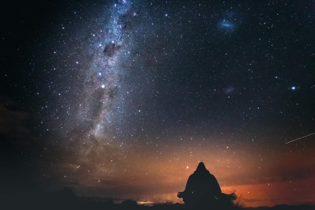 A lone figure sits under a starry sky with the Milky Way visible, capturing the beauty of a New Zealand night.