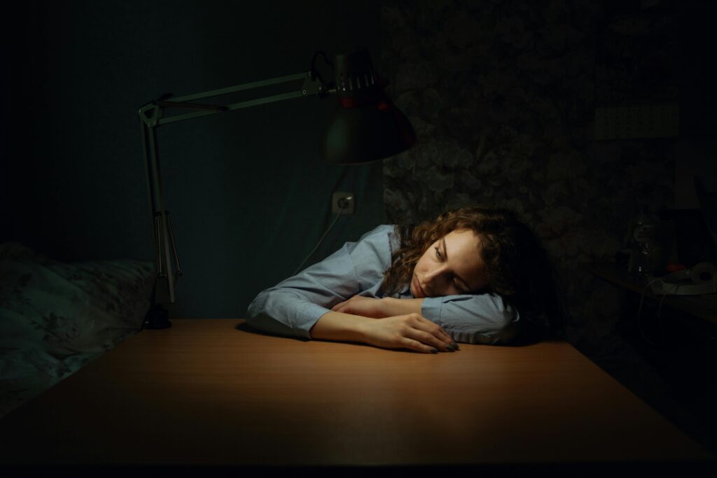 A woman in a dimly lit room rests her head on a desk, illuminated by a desk lamp.